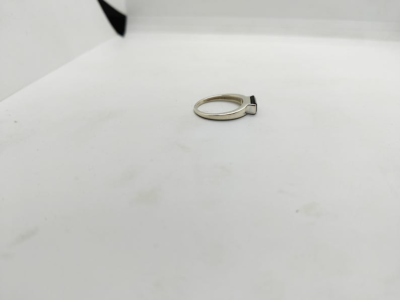May include: A silver ring featuring a rectangular black gemstone. The ring is displayed on a white surface. The band is slender and polished. The black stone is small and square.
