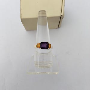 May include: A gold-toned ring with a rectangular, deep purple center stone and two smaller, orange-yellow side stones. The ring is displayed on a clear acrylic stand, with a beige box in the background.