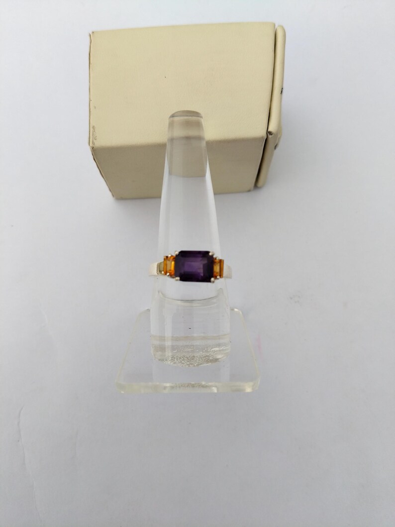 May include: A gold-toned ring with a rectangular, deep purple center stone and two smaller, orange-yellow side stones. The ring is displayed on a clear acrylic stand, with a beige box in the background.