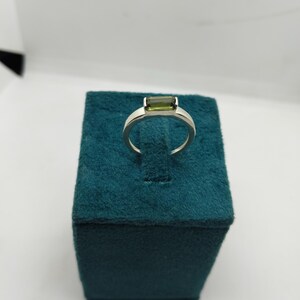 May include: A silver ring featuring a rectangular, olive-green gemstone. The ring is presented on a teal velvet jewellery box, set against a white backdrop. The gemstone is the focal point, showcasing a clean, modern design.