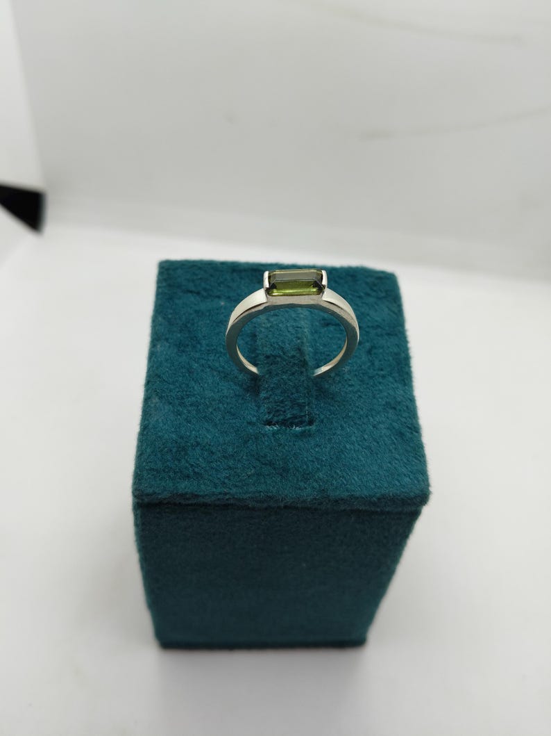 May include: A silver ring featuring a rectangular, olive-green gemstone. The ring is presented on a teal velvet jewellery box, set against a white backdrop. The gemstone is the focal point, showcasing a clean, modern design.