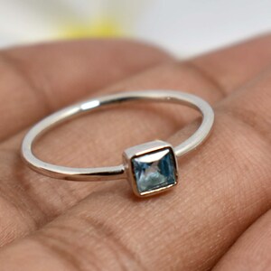 Könnte beinhalten: Ein silberner Ring mit einem quadratischen, hellblauen Edelstein. Das Band ist dünn und poliert, und der Edelstein ist in einer quadratischen Fassung gefasst. Der Ring wird auf einer Hand gezeigt.