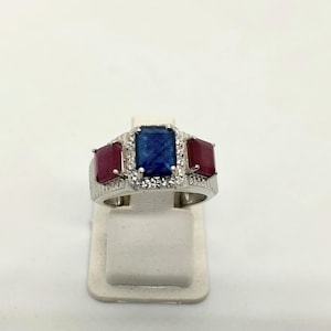 Könnte beinhalten: Ein silberfarbener Ring mit einem zentralen rechteckigen blauen Edelstein, flankiert von zwei kleineren rechteckigen roten Edelsteinen. Der Mittelstein ist von einem Kranz aus kleinen klaren Steinen umgeben, der Ring hat eine texturierte Oberfläche.