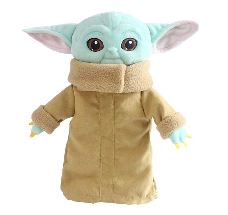 Puede incluir: Un juguete de peluche del personaje Baby Yoda de la serie de Star Wars The Mandalorian. El juguete lleva un abrigo marr&oacute;n con un cuello de piel y tiene grandes ojos marrones.