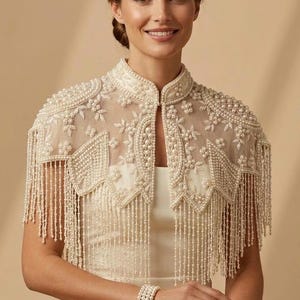 May include: Ivory-colored beaded cape with a high collar and fringe detailing. The cape is adorned with intricate pearl and bead embellishments, creating a delicate and elegant design. The cape is worn over a matching ivory dress, and a pearl bracelet is visible.