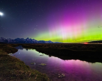 May include: A vibrant green and purple aurora borealis display over a mountain range and a still body of water. The moon is visible in the upper left corner of the image.