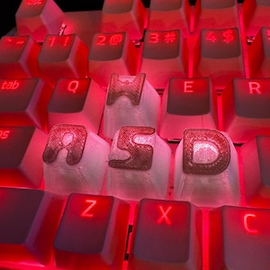 May include: Close-up of a computer keyboard with red backlighting. The keys are white with red illuminated letters and symbols. The keys "W", "A", "S", and "D" are highlighted with a unique design.
