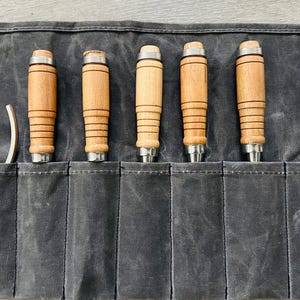 May include: A set of eight wood carving tools, including a curved gouge, are rolled up in a gray canvas tool roll. The tools have wooden handles and metal blades. The tool roll has a label that reads "Chisel Variation 1/2".