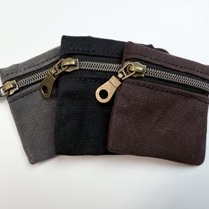 May include: Three small zippered pouches made of canvas fabric. The pouches are gray, black, and brown. Each pouch has a zipper closure and a small metal loop.
