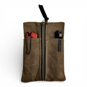 May include: A brown canvas tool roll with a black zipper and a black strap. The tool roll has two pockets, one with a red object and a black clip, and the other with a black object and a black clip.