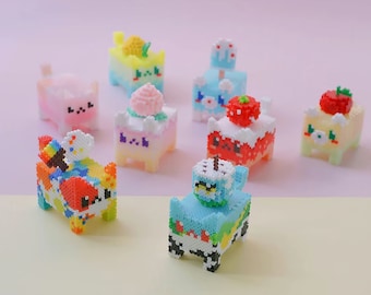 May include: A collection of colorful, pixelated cat figurines with different toppings, including a strawberry, a pineapple, a cloud, and a swirl of frosting. The figurines are made of small beads and are arranged on a pink and yellow surface.