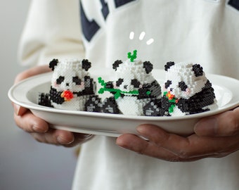 May include: Three white and black panda bead sprites on a white oval plate. The pandas are holding flowers and leaves.