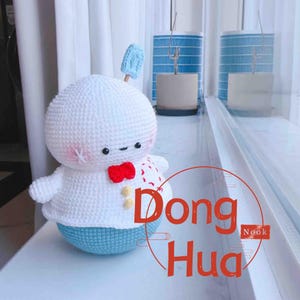 Puede incluir: Muñeca blanca de ganchillo hecha a mano con cabeza redonda, brazos pequeños y base azul. La muñeca tiene una pajarita roja y un pequeño accesorio azul en la parte superior de la cabeza. El texto "Dong Hua" está en la imagen.