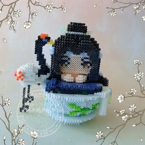 May include: A beaded figurine of a person with black hair and a blue and white outfit sitting in a blue and white teacup with a green leaf design. A white beaded crane with an orange beak is next to the teacup.