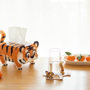 Pode incluir: Um dispensador de caixas de lenços de papel em forma de tigre laranja e preto. O tigre tem um rosto amigável e uma cauda listrada. Um copo de água, um descanso de copo de tigre e um prato de tangerinas estão em uma superfície de madeira.