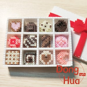 May include: A white box of handmade chocolates, each crafted from small, colorful beads. The chocolates feature various shapes and colors, including pink hearts, brown swirls, and white and brown patterns. A gift box with a red bow is in the background.
