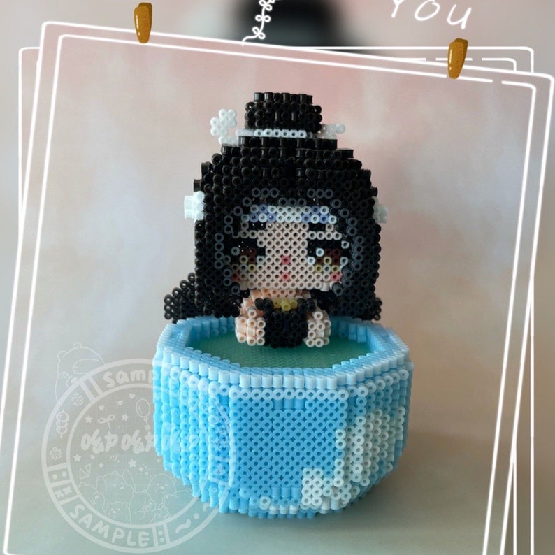 May include: A blue and white bead sprite of a person with black hair and a black swimsuit sitting in a blue pool. The bead sprite is surrounded by a white border.