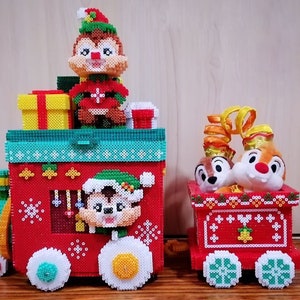 Könnte beinhalten: Zwei Spielzeugzüge zum Thema Weihnachten. Der größere Zug ist rot mit einem grünen Dach und zeigt einen Chipmunk-Charakter in einem Elfen-Outfit. Der kleinere Zug ist ebenfalls rot und hat zwei Chipmunk-Charaktere im Inneren. Beide Züge haben weiße und grüne Räder.