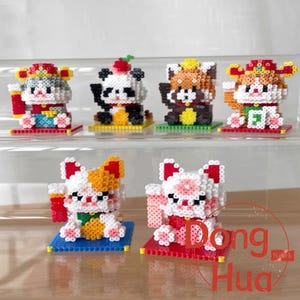 May include: A selection of colourful, pixelated figurines, including lucky cats and panda bears. The figurines are made of small, interlocking blocks and sit on coloured bases. The figurines have detailed features and are arranged on a clear shelf.