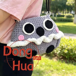 May include: A gray crocheted bag shaped like a creature with large white and black eyes, a black nose, and pink cheeks. It features a wavy white trim across the front and two black circular feet at the bottom. The bag has a black strap and text reading Dong Hua Nook.