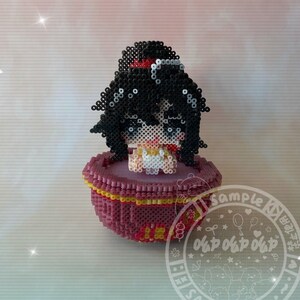 May include: A small, colorful, 3D bead art figure of a girl with black hair and a white dress sitting in a red and yellow bowl. The figure is made of perler beads.