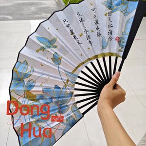 Può includere: Un ventaglio con manico nero e stecche bianche e nere. La superficie del ventaglio presenta illustrazioni di fiori di loto blu con accenti dorati e calligrafia cinese. Il ventaglio è tenuto aperto, mostrando il disegno.