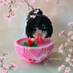 May include: A pink and white beaded figurine of a girl with black hair, wearing a pink dress and holding a pink flower. The figurine is sitting on a pink and white beaded base. The background is a blurry image of pink flowers.