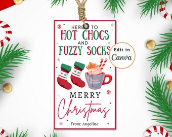 Pode incluir: Uma etiqueta de presente de Natal vermelha e verde com uma ilustração festiva de uma caneca de chocolate quente, meias felpudas e o texto "Here to hot chocs and fuzzy socks Merry Christmas From: Angelina".