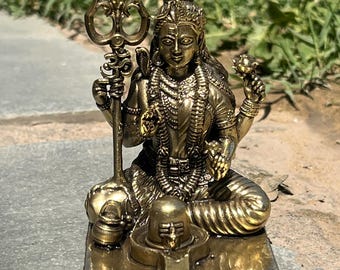 May include: A detailed brass statue of a seated deity, holding a trident and a lotus flower. The figure is adorned with intricate jewelry and a crown. The statue sits atop a rectangular base with a detailed design. The overall color is a warm, antique gold.