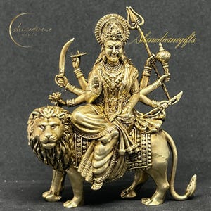 May include: A gold-colored statue of a multi-armed deity riding a lion. The deity wears ornate jewelry and holds various weapons. The statue has the text "Shinedivinegifts" in a cursive font.