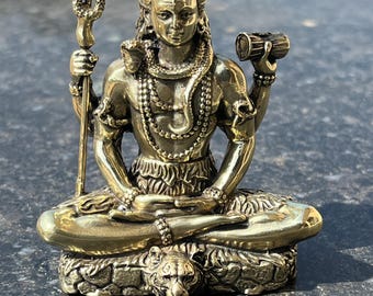 May include: A detailed brass statue of a seated deity, possibly Shiva, with multiple arms and intricate jewelry. The figure holds a trident and a small drum. The statue is on a base with a tiger head. The overall color is a warm, antique gold.