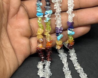 May include: A close-up of several necklaces made of colorful, small, irregular gemstones. The gemstones are arranged in a rainbow pattern, with colors including purple, blue, green, yellow, orange, and red. The necklaces are held against a dark background.