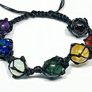 Puede incluir: Una pulsera de macramé con siete cuentas de piedras preciosas de colores, cada una encerrada en una red negra tejida. Las piedras preciosas incluyen amatista, lapislázuli, jade, citrino, cornalina y una piedra de color rojo oscuro. La pulsera es ajustable con un cordón negro.