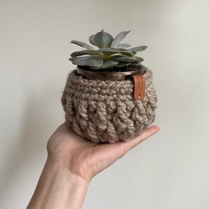 Op de afbeelding: Een kleine vetplant in een koperkleurige pot, omhuld door een handgehaakte, lichtbruine hoes. Een klein leren label is aan de hoes bevestigd. De plant heeft lichtgroene bladeren.