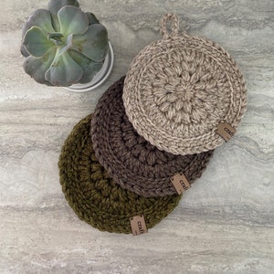 May include: Three handmade crochet pot holders in olive green, brown, and tan. Each round pot holder has a loop for hanging and a small tag that reads "craft". A succulent plant in a white pot is in the background.