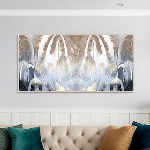May include: A large abstract painting with a symmetrical design of white and gold feathers on a blue background. The painting is framed in a thin, gold frame and is hanging on a white wall above a beige sofa with cushions.