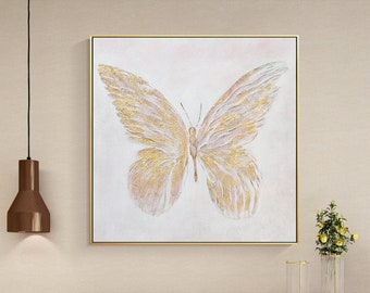 May include: A white cabinet with gold accents and a geometric pattern. The cabinet has two doors and is sitting on a light brown floor. A framed painting of a gold butterfly on a pink background is hanging on the wall above the cabinet.