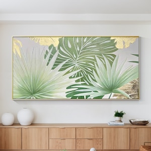 Puede incluir: Una gran pintura sobre lienzo enmarcada con un diseño de hojas tropicales verdes y doradas. La pintura está colgada en una pared blanca sobre un armario de madera.