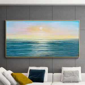 May include: A painting of a seascape with a blue ocean and a bright yellow sun in the sky. The painting is framed in gold and hangs on a gray wall above a white couch.