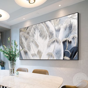 Puede incluir: Pintura abstracta en tonos blancos, grises y dorados. La pintura presenta un primer plano de pétalos superpuestos, creando un efecto texturizado y en capas. La pintura está enmarcada en un marco negro y cuelga de una pared blanca.