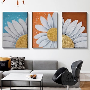 Pode incluir: Três pinturas abstratas de uma margarida branca com um centro dourado. As margaridas são pintadas em um fundo azul, laranja e marrom.