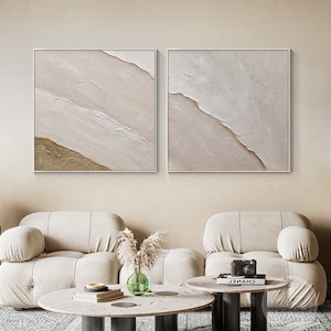 May include: Two abstract paintings with a beige and white color scheme. The paintings are framed and hung on a beige wall above a white couch.