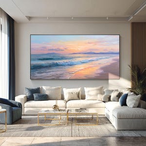 Op de afbeelding: Grote rechthoekige canvas kunst die een strandscène bij zonsondergang afbeeldt. Het kunstwerk toont een levendige lucht met tinten van oranje, roze en blauw, die weerspiegeld worden in de oceaangolven en het natte zand. Het schilderij is zwart omlijst.