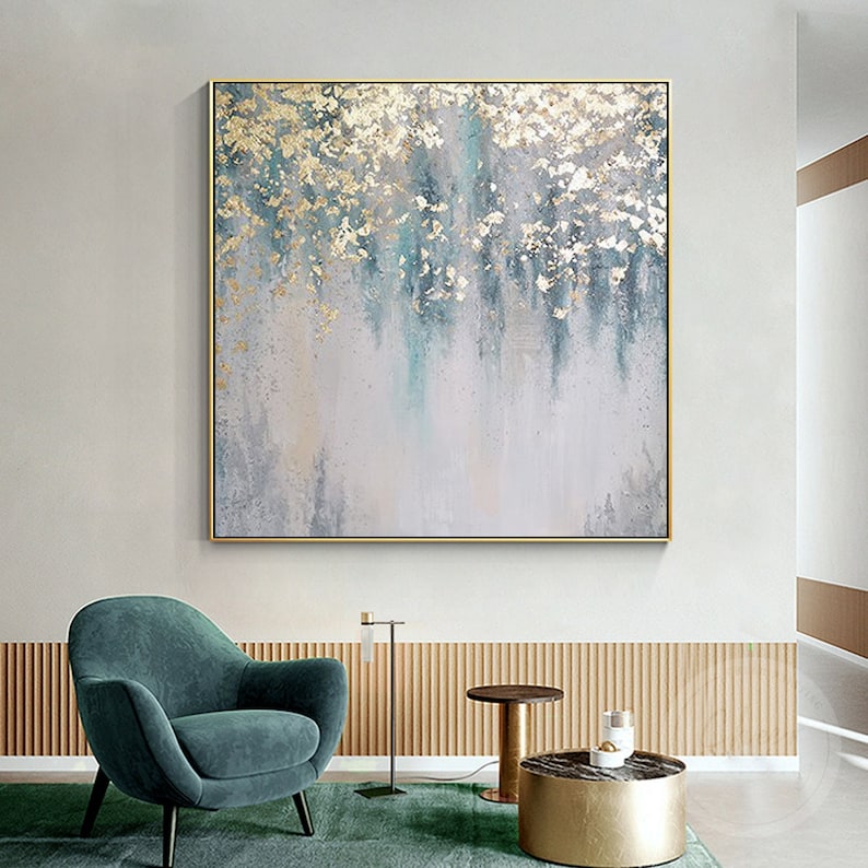 May include: Abstract painting with gold and blue accents on a white canvas. The painting is framed in a gold frame and is hanging on a white wall.