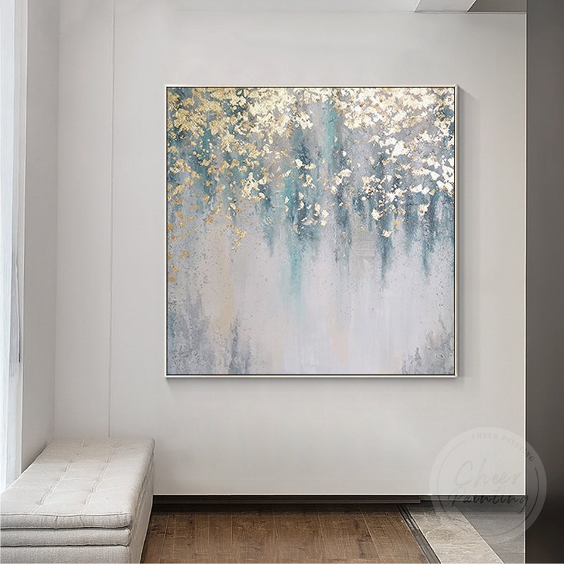 Op de afbeelding: Abstract schilderij met een witte achtergrond en gouden en blauwe accenten. Het schilderij heeft een textuur en is ingelijst in een dun, zilveren lijst.