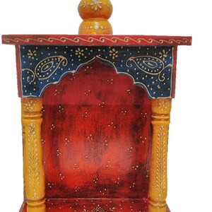 May include: A hand-painted wooden temple in a vibrant colour scheme. The structure features a red interior, blue accents with paisley designs, and yellow pillars. The top is adorned with a yellow finial, and the base is red with a white floral pattern.