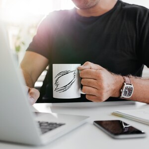 May include: A person sitting at a desk with a laptop, a cell phone, and a white mug with a black doodle design. The person is wearing a black t-shirt and a watch.