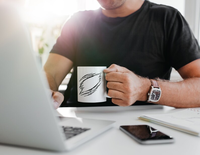May include: A person sitting at a desk with a laptop, a cell phone, and a white mug with a black doodle design. The person is wearing a black t-shirt and a watch.