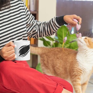 May include: A person wearing a black and white striped shirt and red pants sits on the floor with a white mug with a black design. A ginger and white cat plays with a pink and purple toy.