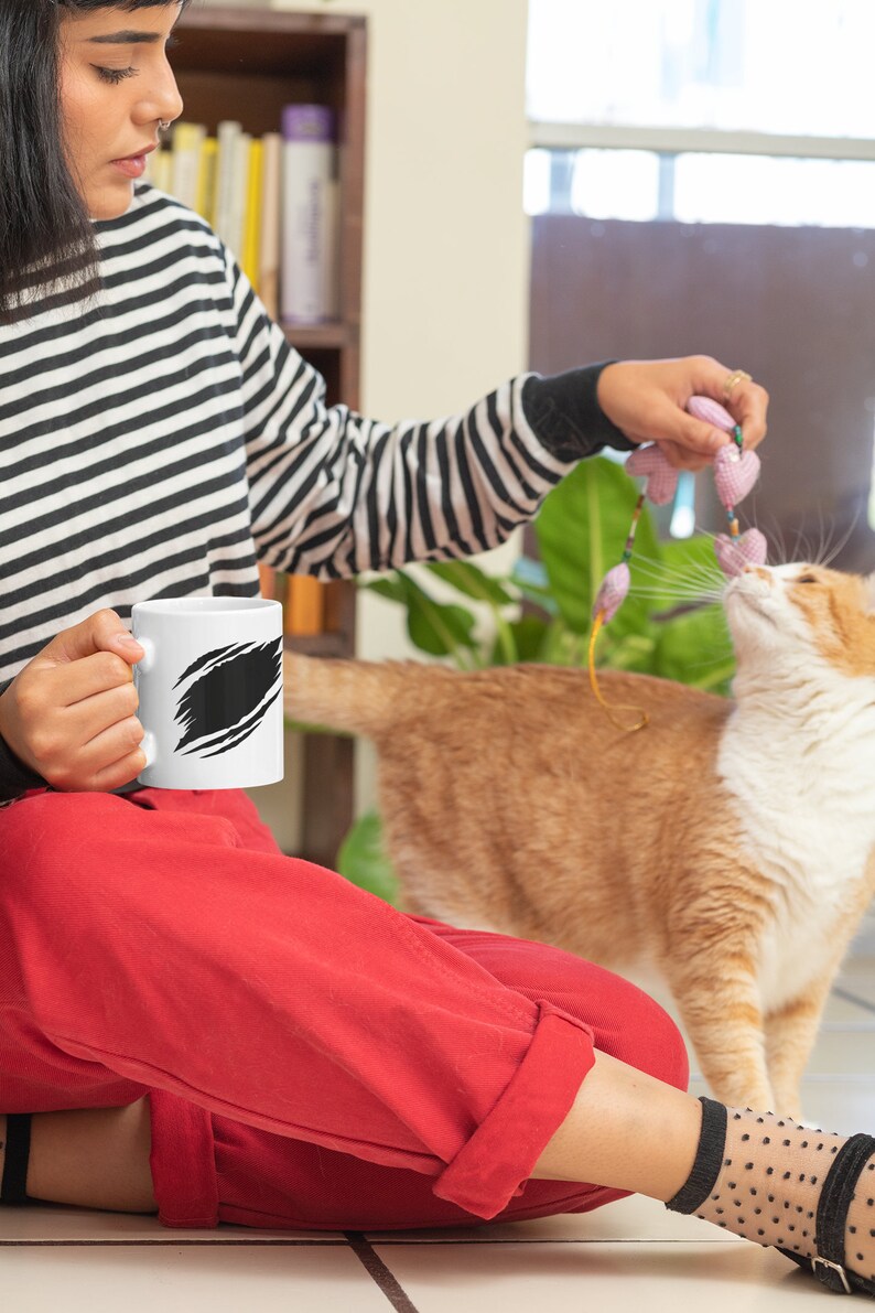 May include: A person wearing a black and white striped shirt and red pants sits on the floor with a white mug with a black design. A ginger and white cat plays with a pink and purple toy.